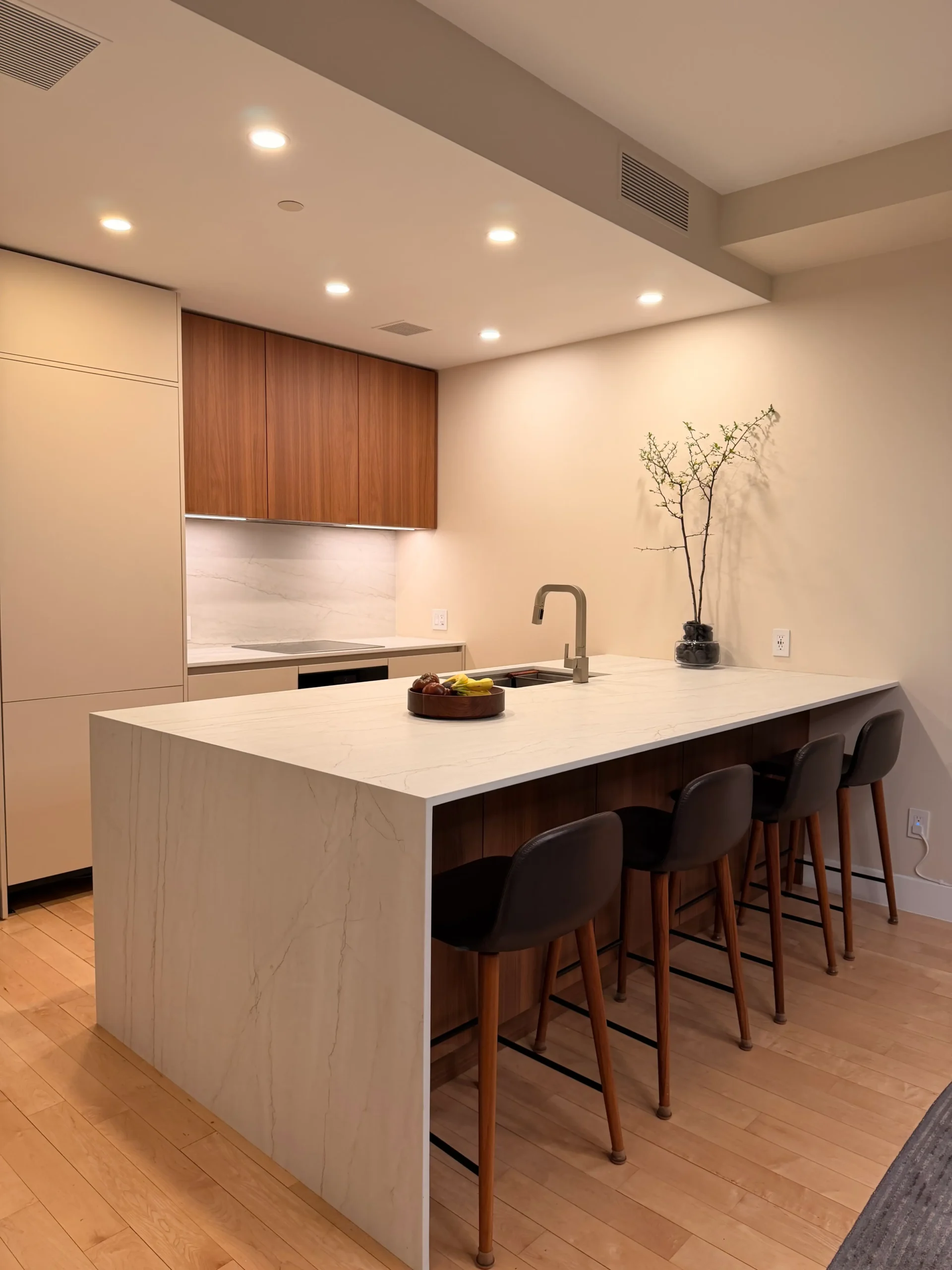 Modern kitchen with a marble island and bar seating along a light wood floor, warm recessed lighting, and wooden cabinets.